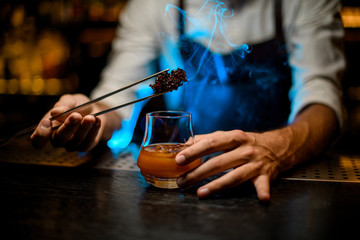 Male barman adding chilled melting caramel with twezzers to the cocktail glass with ice cubes under blue lights