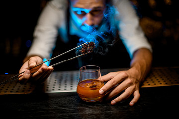 Professional bartender adding chilled melting caramel with twezzers to the cocktail with ice cubes under blue lights