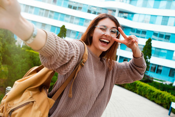 Fototapeta premium Young girl take selfie from hands with phone on summer spring city street. Urban life concept.Closeup selfie-portrait student of attractive girl in sunglasses with long hairstyle and snow-white smile 