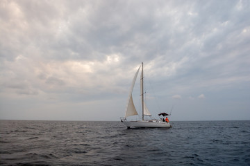 Fototapeta premium White sail boat on the grey calm water of sea or ocean during cloudy, windy and grey day with yacht team on board 