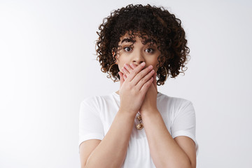 Fototapeta na wymiar Awkward cute curly-haired silly alluring girl slept secret feel sorry say oops cover mouth palms pressed lips say something inappropriate, standing white background worried stunned