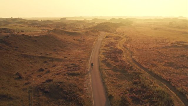 Aerial shot of a young woman cyclist riding a bike on a bike path among sand dunes at golden sunrise in the Texel island in The Netherlands