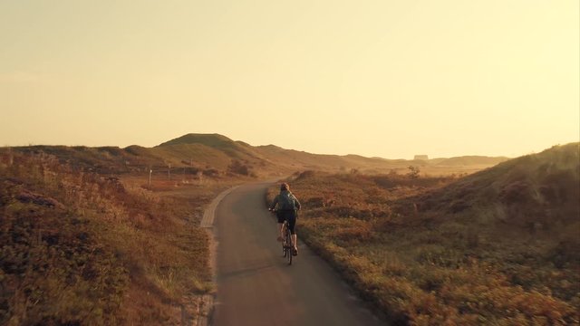 Tracking aerial shot of a young woman cyclist riding a bike on a bike path among sand dunes at golden sunrise in the unique landscape of the Texel island in The Netherlands