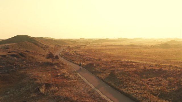 Tracking pull back aerial shot of a young woman cyclist riding a bike on a bike path among sand dunes at golden sunrise in the unique landscape of the Texel island in The Netherlands