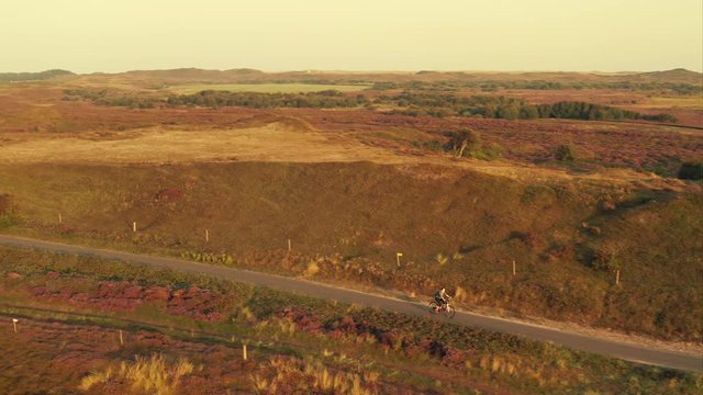 Tracking aerial shot of a young woman cyclist riding a bike on a bike path among sand dunes at golden sunrise in the unique landscape of the Texel island in The Netherlands