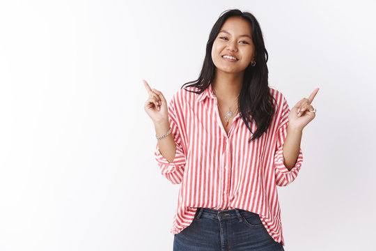 Girl recommends to items both good. Portrait of happy good-looking asian young female shop assitant pointing up right and left smiling cute at camera as giving suggestions what buy over white wall