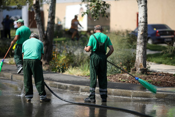Street sweeper cleaning city sidewalk with water from a hose and a plastic broom - blue collar jobs