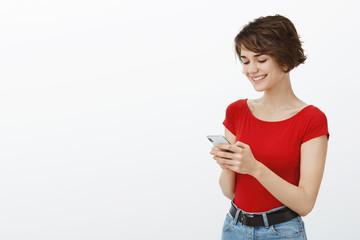 Friendly sociable good-looking brunette female short haircut hold smartphone smiling happy delighted screen display texting using app messaging friends stand white background grin screen