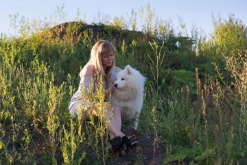 Horizontal frontal view of pretty blond young woman sitting on slope covered in wildflowers petting...