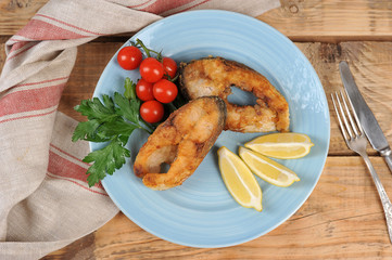 Carp steaks on a plate. Lemon slices, cherry tomatoes and parsley complete the fried fish dish. Wood background. View from above.