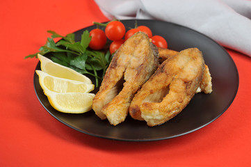 Carp steaks on a black plate. Lemon slices, cherry tomatoes and parsley complete the fried fish dish. Red background. Close-up.