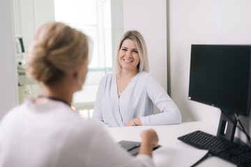 Young blonde woman at doctor reception in white dressing gown. Therapist female sitting at table and asks the patient questions