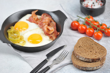Fried eggs with bacon in a pan. Slices of dark bread, cutlery, cherry tomatoes and spices complete the composition. Close-up. Light background.