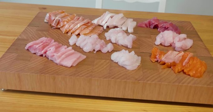 Slow pan around a beautiful presentation of fresh sliced sashimi fish on a wooden board sitting in the middle of wooden table. Fish includes raw tuna, salmon, kingfish and snapper