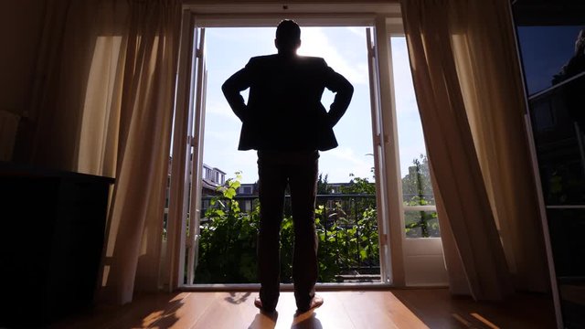 Handsome man open curtains and stay against full height window, look to sunny outdoors. Bedroom at second floor cosy town house, typical living at European country. Guy wear casual suit