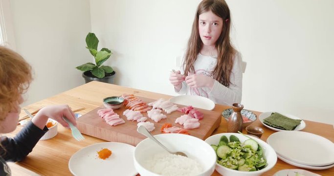 young girl holding beginner chopsticks about to start eating fresh raw sashimi while a little boy eats orange fish row - fish eggs. The raw fish is laid out on a board in the center of the table