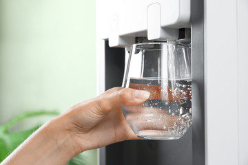 Woman filling glass from water cooler indoors, closeup. Refreshing drink