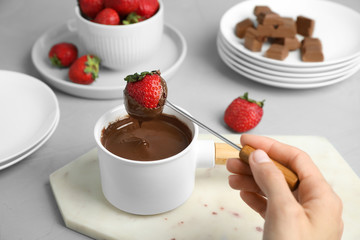 Woman dipping strawberry into fondue pot with chocolate at grey table, closeup