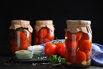 Pickled tomatoes in glass jars and products on black wooden table