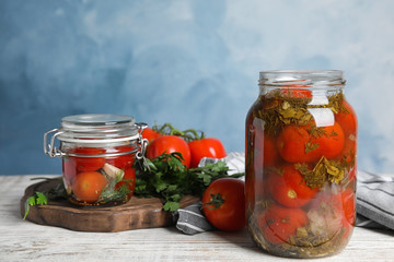Pickled tomatoes in glass jars and products on white wooden table against blue background