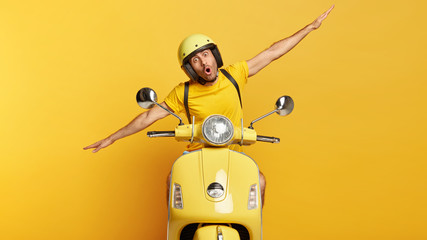 Impressed young male motorcyclist spreads hands sideways, drives motorbike, enjoys extreme drive, wears yellow t shirt, helmet, isolated over yellow background. Monochrome shot. Transport and people