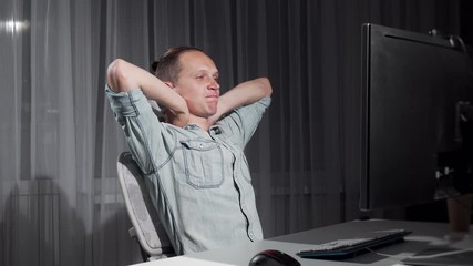 Happy man finishing work on his computer late at night. Handsome young male freelancer stretching in his chair after finishing his project. Success, studying concept