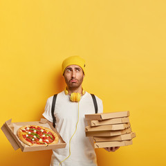 Image of dissatisfied delivery man holds pile of cardboard boxes, shows tasty cheese pizza, has sad expression, wears yellow hat and white t shirt, isolated on yellow background, empty space