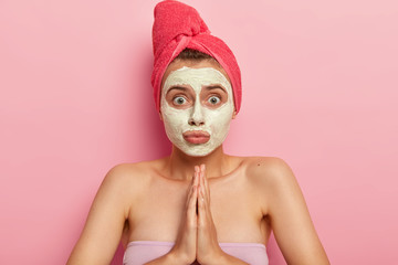 Photo of surprised young woman keeps palms together in praying gesture, has helpess puzzled face expression, wears nourishing facial mask for reducing wrinkles, uses homemade cosmetic ingredients