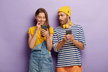 Millenial girl looks positively at smartphone device, shocked puzzled guy with cellular, stand closely to each other against purple background. Youth with modern technologies. Addicted couple