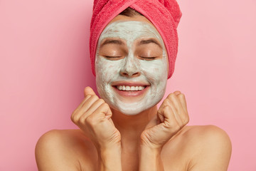 Close up portrait of happy female model with facial clay mask on face, clenches fists, receives good news, keeps eyes closed, wears pink towel on head, stands bare shoulders has rejuvenation procedure