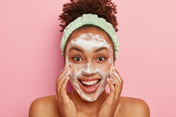 Image of glad dark skinned young female model has crisp hair, washes face with soap, cleans from pores, has problematic teenage skin, stands naked, wears headband, isolated over pink background - obrazy, fototapety, plakaty
