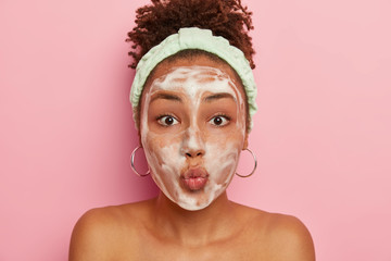 Teenage girl with dark skin has problematic skin, applies foam to wetter face, keeps lips folded at camera, poses topless, wears turquoise headband and earrings stands topless against pink studio wall - obrazy, fototapety, plakaty