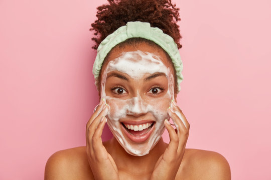 Image of glad dark skinned young female model has crisp hair, washes face with soap, cleans from pores, has problematic teenage skin, stands naked, wears headband, isolated over pink background