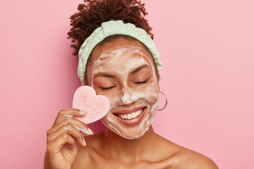 Isolated shot of positive dark skinned woman with Afro combed hair, wears headband, cares about...