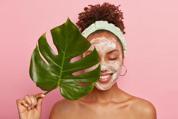Portrait of happy African American woman covers half of face with green leaf, cleans face, washes with bubble soap, stands topless, cares aboout her beauty and body, isolated over pink background