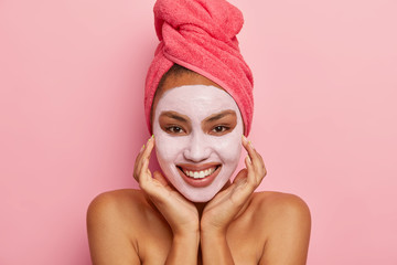 Isolated shot of pretty woman touches face gently, wears facial mask for refreshing skin, has healthy complexion, wears pink towel, has cosmetic treatments, stands indoor. Femimity, spa, relaxation