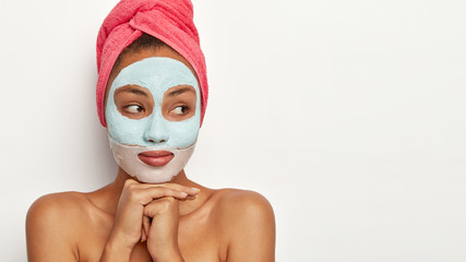 Cropped image of beautiful pensive young woman concentrated aside, keeps hands under chin, looks away, wears clay mask, towel on wet hair, cleans face from pores, models against white background