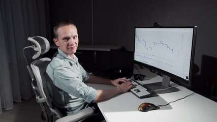 Young male IT programmer smiling to the camera while working on his project. Charming young freelancer working online on his computer late at night. Work, career concept