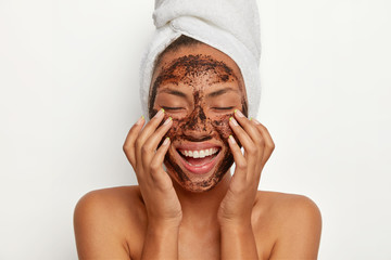 Portrait of cheerful smiling dark skinned woman applies natural coffee mask, makes circular motions with hands and massages skin, stimulates facial blood supply, wears wrapped towel on head.