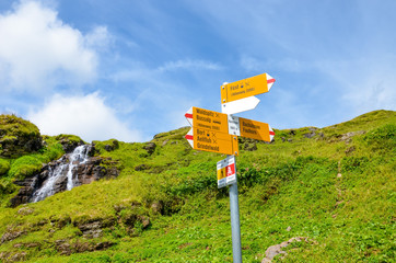 Yellow tourist sign in Bachlager, Switzerland giving distances and directions to hikers in the Swiss Alps. Popular hiking paths by Grindelwald leading to Bachalpsee. Alpine stream in background