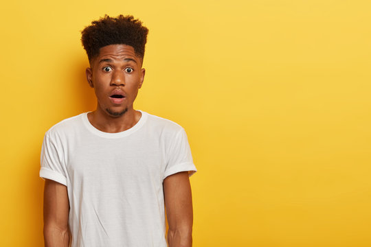 Concerned panicking man stressed by puzzled situation, has bated breath, healthy dark skin and curly hairstyle, little beard, poses against yellow background with copy space for your advertisement