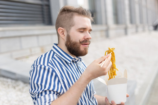 Close-up Of An Amusing Young Hipster Guy Eating Chinese Noodles With Wooden Chopsticks Sitting In A Park Outside On A Warm Summer Day. The Concept Of Rest And Snacking On The Street.