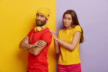 Displeased guy has turns back to girlfriend, keeps hands crossed, doesnt speak to her, wears yellow hat and casual red t shirt, unhappy woman touches shoulder of boyfriend. Couple, relationship