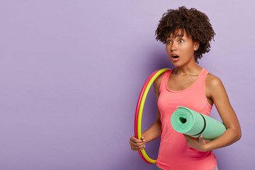 Fototapeta na wymiar Surprised ethnic woman with curly hairstyle, carries hula hoop, has training for loosing weight, leads healthy lifestyle, carries karemat, wears casual pink vest, stands over purple wall, empty space