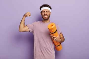 Motivated powerful man stands with fitness mat, enjoys yoga as sport and hobby, raises arm and shows muscle, clenches teeth, wears headband, violet t shirt. Balance your life, lead healthy lifestyle