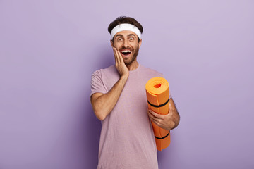 Positive Caucasian man stands with rolled up karemat, touches cheek, wears headband and t shirt,...