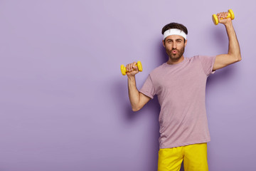 Horizontal shot of handsome unshaven man has workout in gym, trains biceps with sport instructor,...