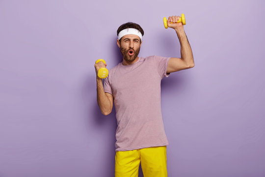 Funny man has fun, exercises with dumbbells, dressed in active wear, motivated for healthy lifestyle, has regular training in morning, isolated over purple background. Athletic guy holds weights