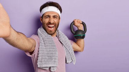 Glad smiling man with toothy smile, wears white headband, lifts heavy weight, takes selfie on unrecognizable device, feels proud of being active sportsman, wears towel on shoulders, headband