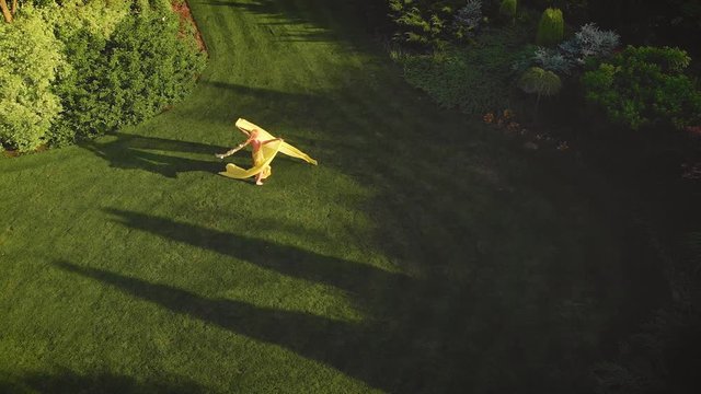 Joyful blonde woman in a bright yellow dress with a train, having fun and spinning in the park. Background green lawn in royal garden. The fabric flies, flutters in the wind. Establishing Aerial View.
